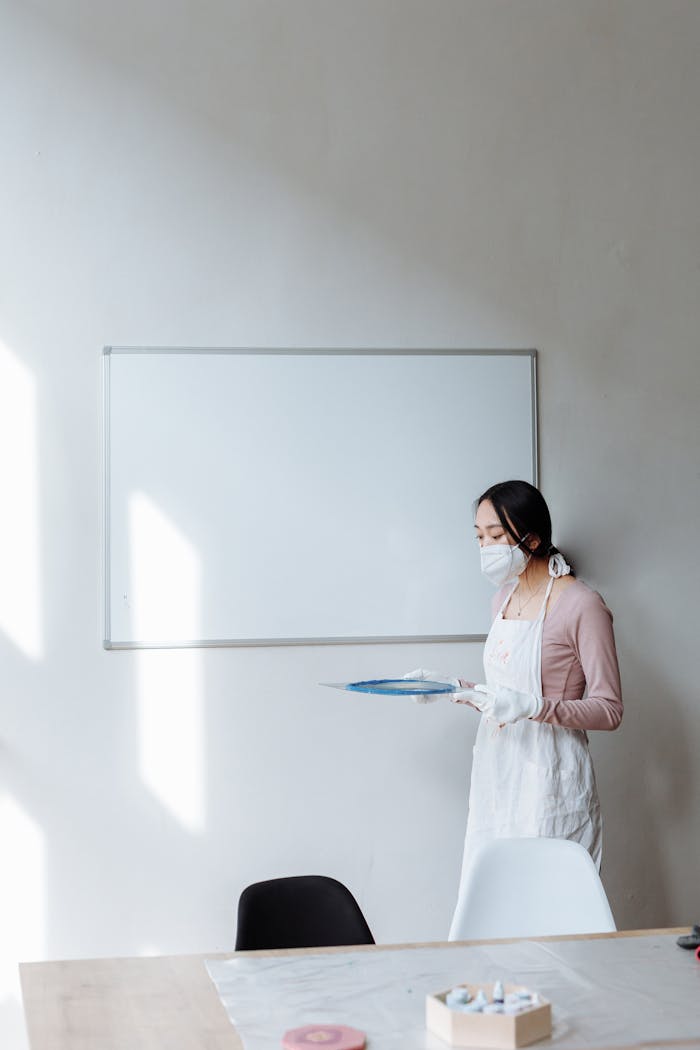Asian woman in apron and mask standing in room with whiteboard, holding items. Indoor setting.